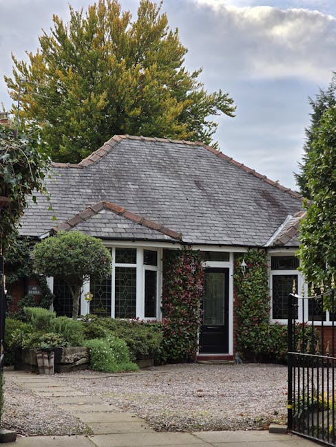 A close-up view of a small front garden in Beckton, featuring a white-framed window with sheer curtains, set within a brick exterior wall. To the left of the window, a hanging basket filled with vibra